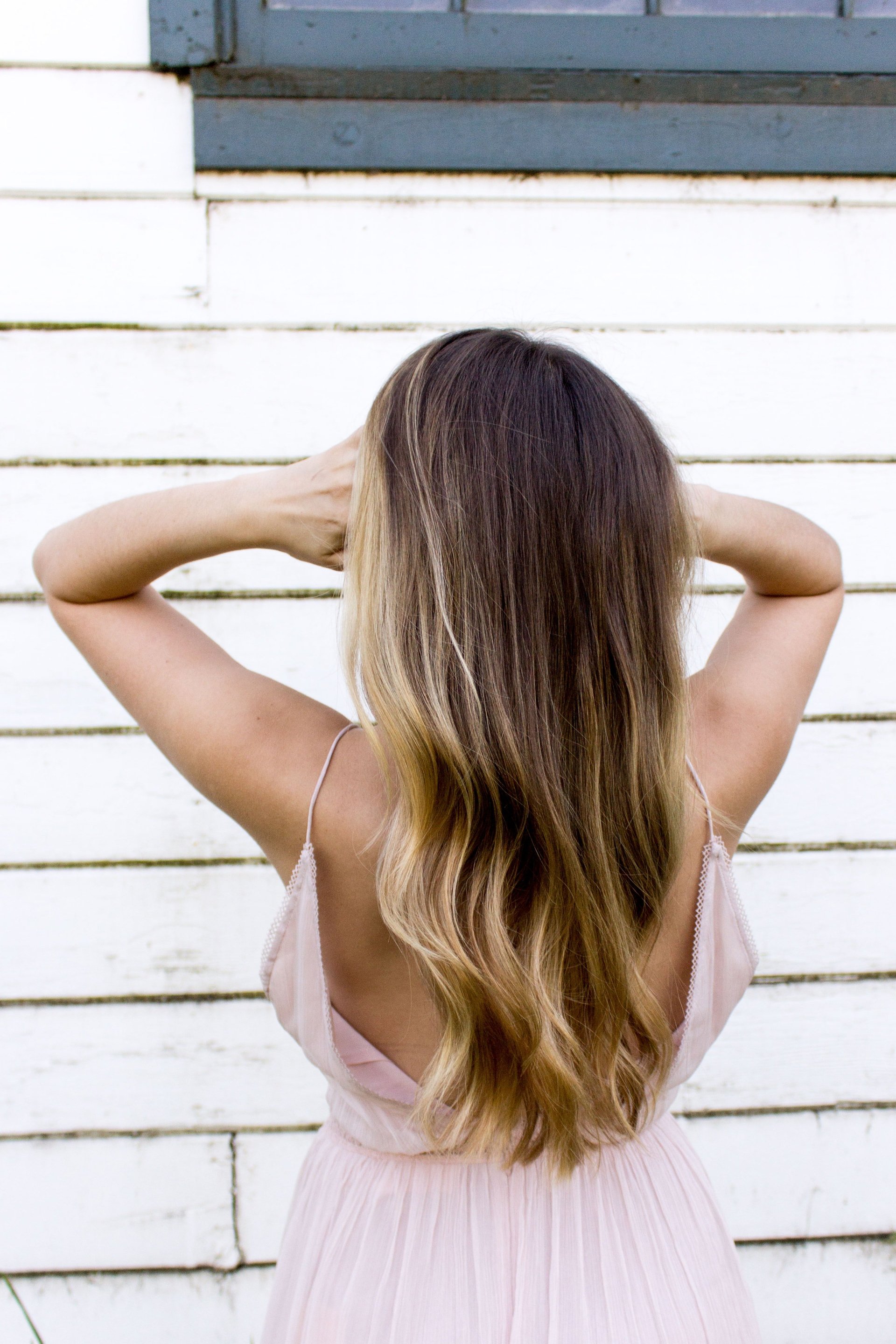 Woman in pink dress, arms raised, against white-painted wall with window. Long hair, ombre effect.